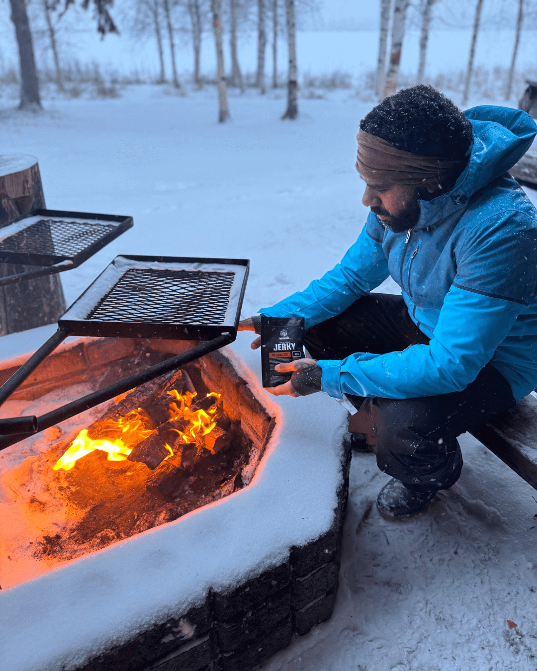 Un homme en tenue hivernale savourant du jerky Greatest Warriors près d'un feu de camp dans un décor enneigé.